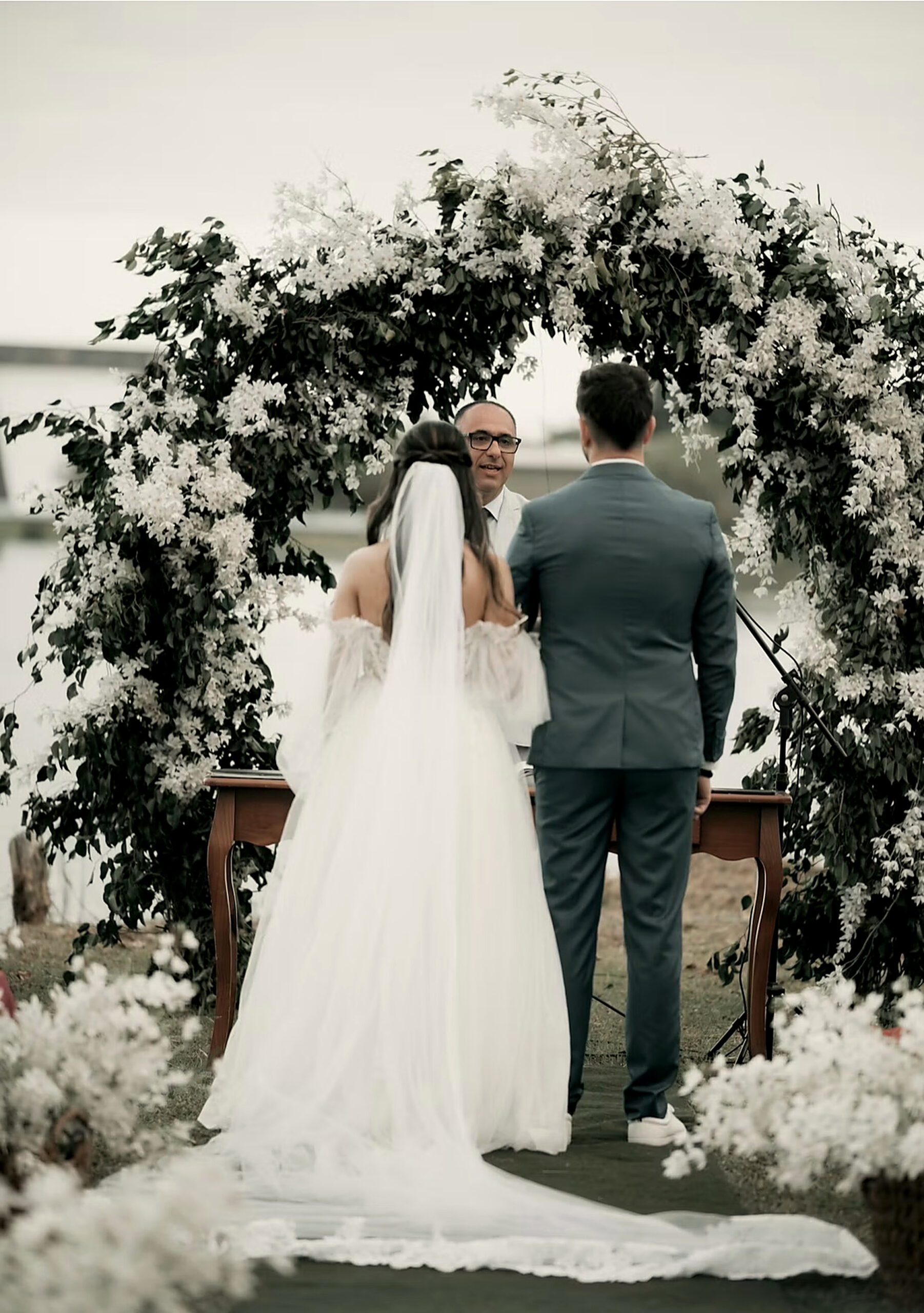 The couple before an arch of white flowers and foliage during a symbolic ceremony by the water, French Riviera