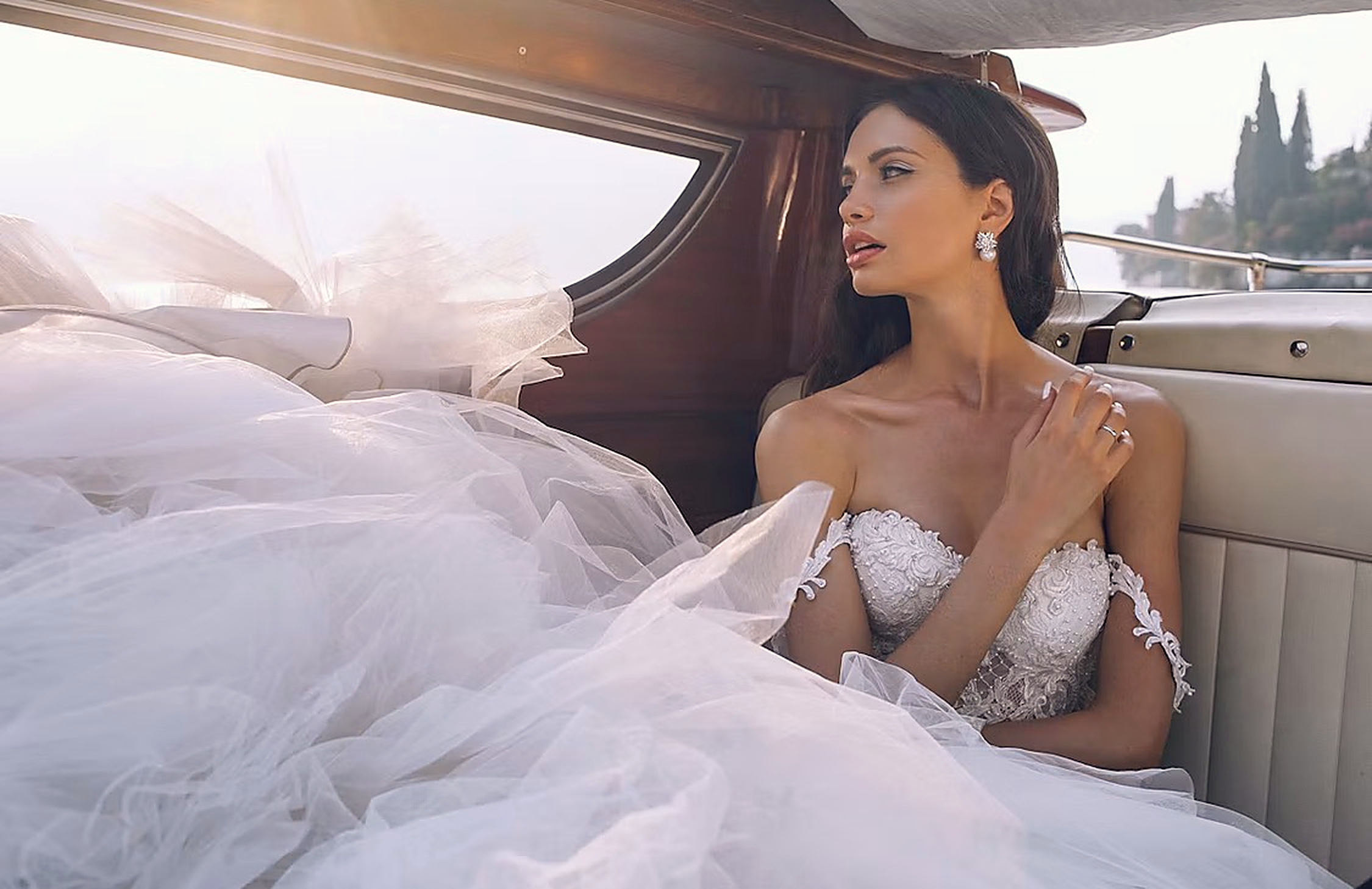 The bride in an embroidered strapless tulle gown aboard a classic varnished wooden boat, cypress trees behind, French Riviera