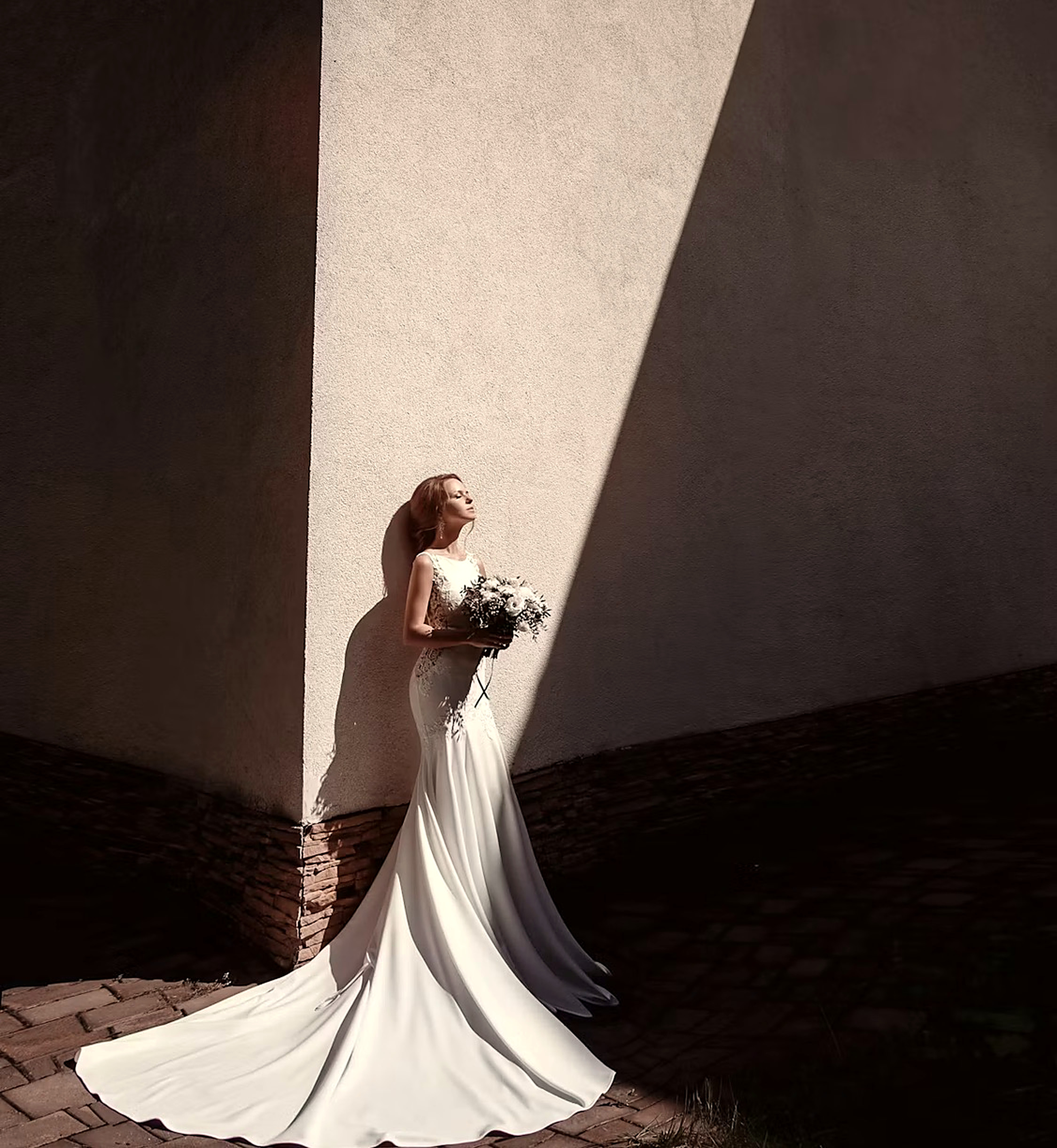 The bride in a mermaid gown with a long train, white bouquet in hand, bathed in sunlight against a stone wall, Saint-Tropez