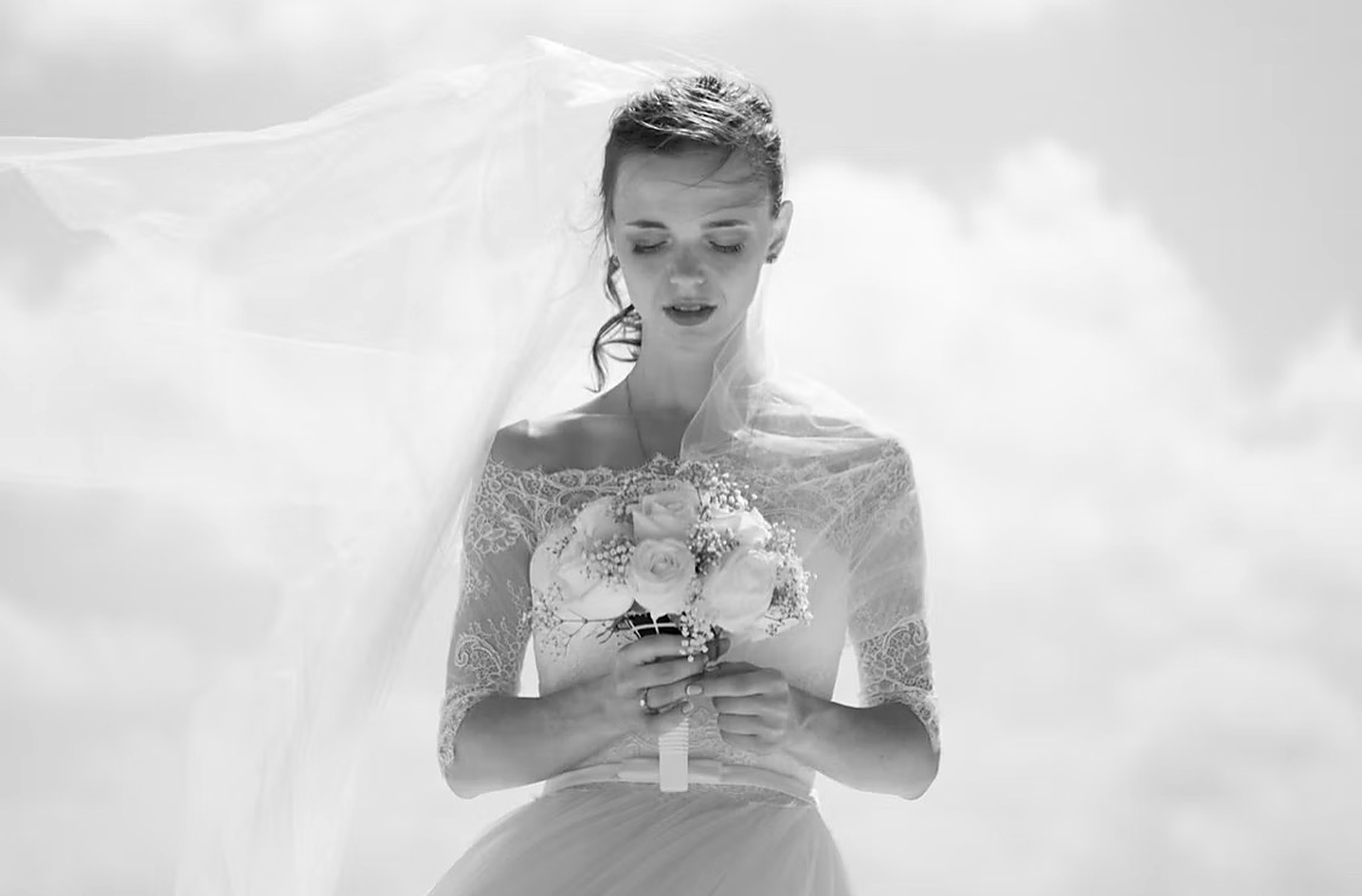 The bride in a lace gown, veil caught by the wind, holding white roses, black and white portrait