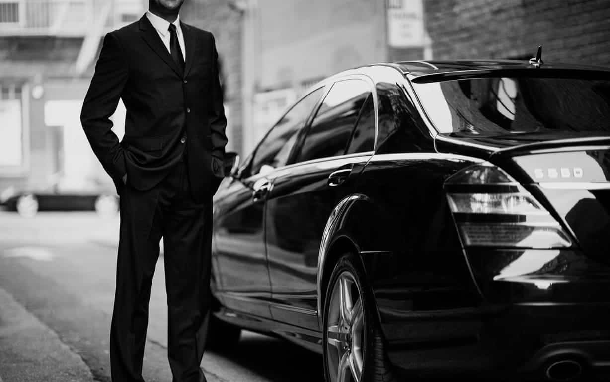 Chauffeur privé en costume devant une berline de luxe, en noir et blanc