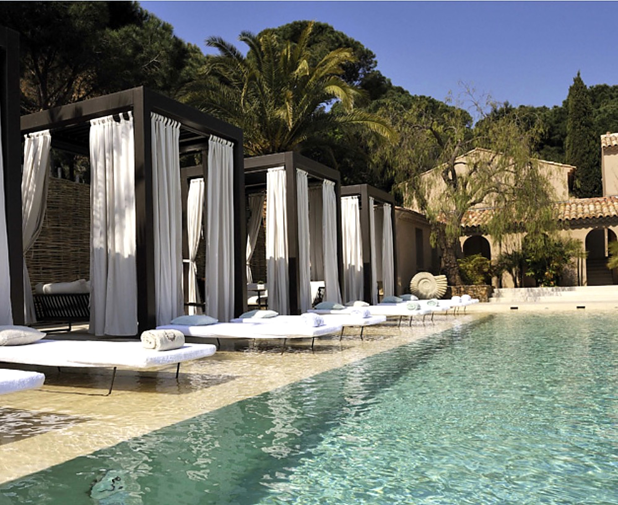 La piscine de l’hôtel Muse à Ramatuelle : lits de plage blancs, cabanas aux rideaux de lin et bastide provençale sous les pins